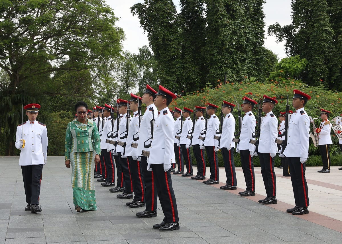 Woman in green dress inspects honor guard in white uniforms with red accents.